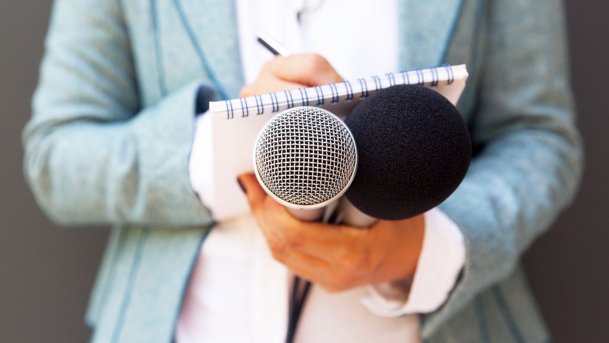 Frontalaufnahme zweier Hände, die zwei Mikrofone halten und auf einem Schreibblock Notizen machen